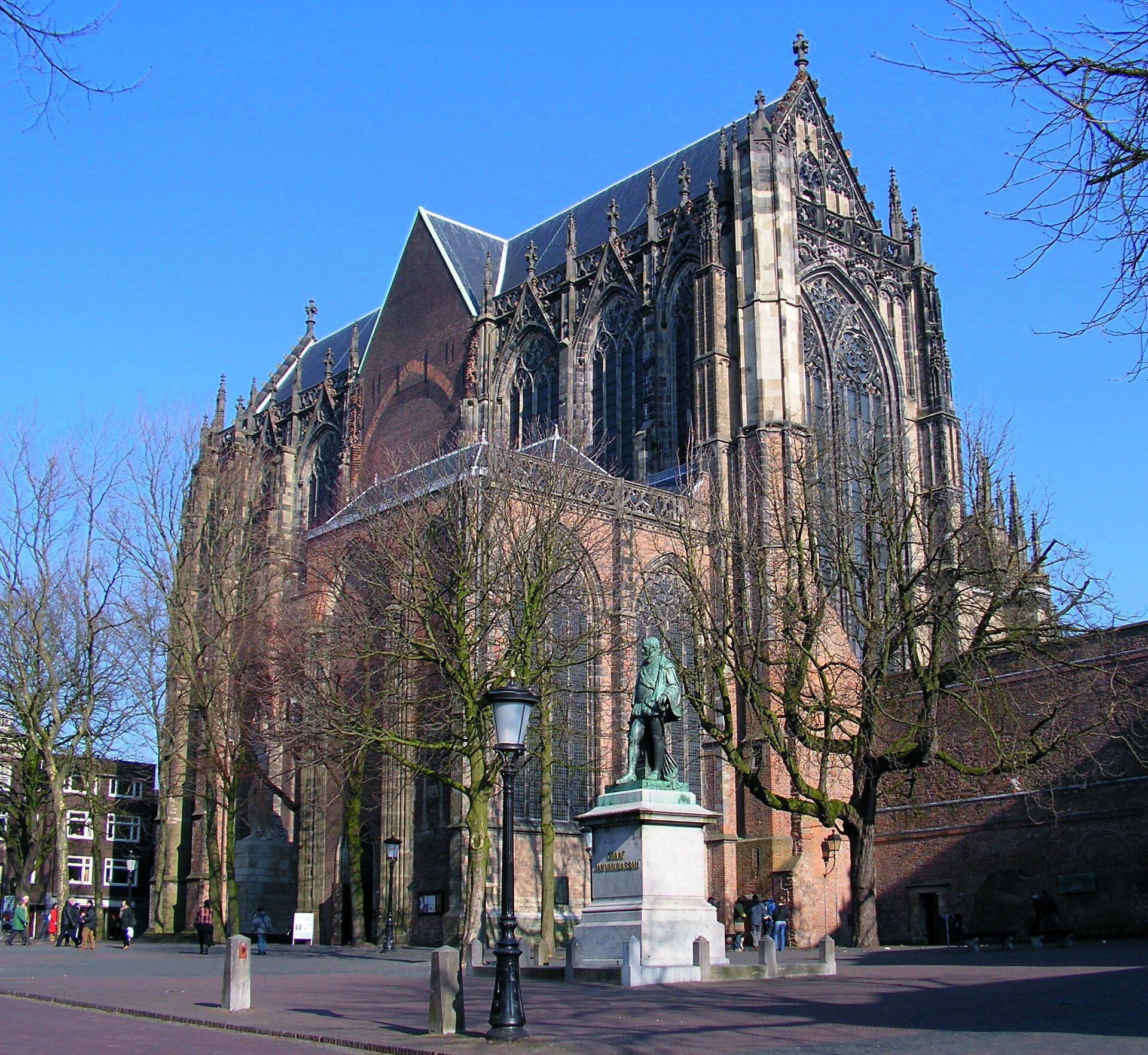 The Gothic Dom Church on Domplein, with a bronze statue in front, bare trees and people walking past.